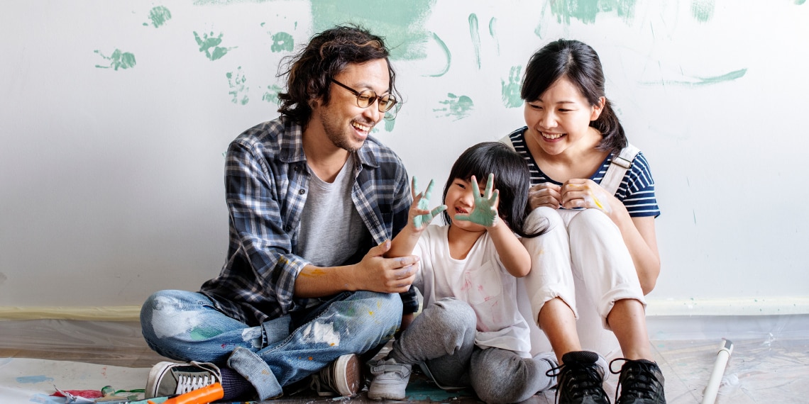 Photo of smiling asian family sitting on floor of house being painted.