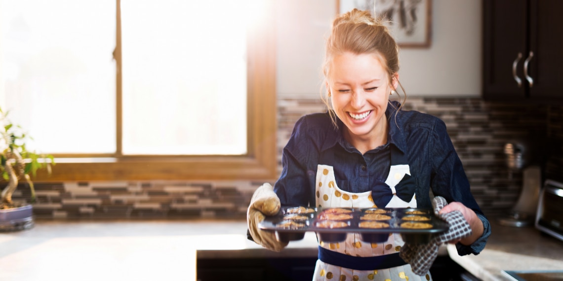 Photo of a woman smiling as she is holding a tray of muffins she just baked.